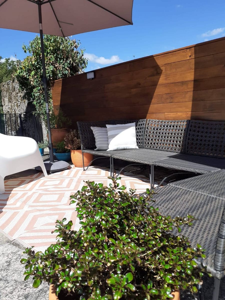 O lounge exterior, com o sofa de vime, o guarda-sol e o muro em madeira.
